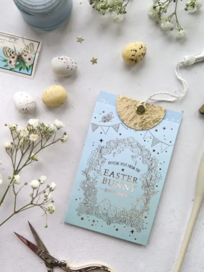 A blue gold foil pouch reads "Official Visit from the Easter Bunny, Delivery Receipt," with a gold half-moon tab and string tie. Speckled mini eggs, a vintage butterfly stamp and gypsophila are scattered around it.