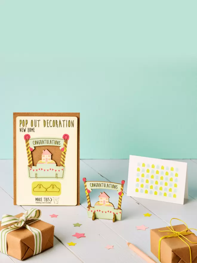 3D new home decoration and house pattern greeting card and brown kraft envelope on top of a grey desk in front of a light green coloured background