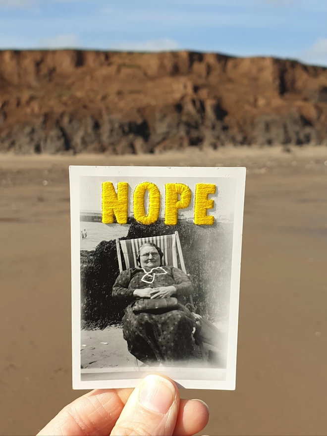 B & W photo of woman with yellow embroidered ‘nope’, held against beach background 