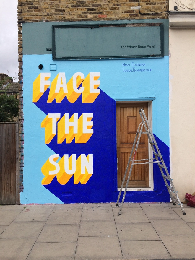 A Survival Techniques street art painting with 3d typography of the words Face The Sun in yellow on a blue wall with a ladder in front