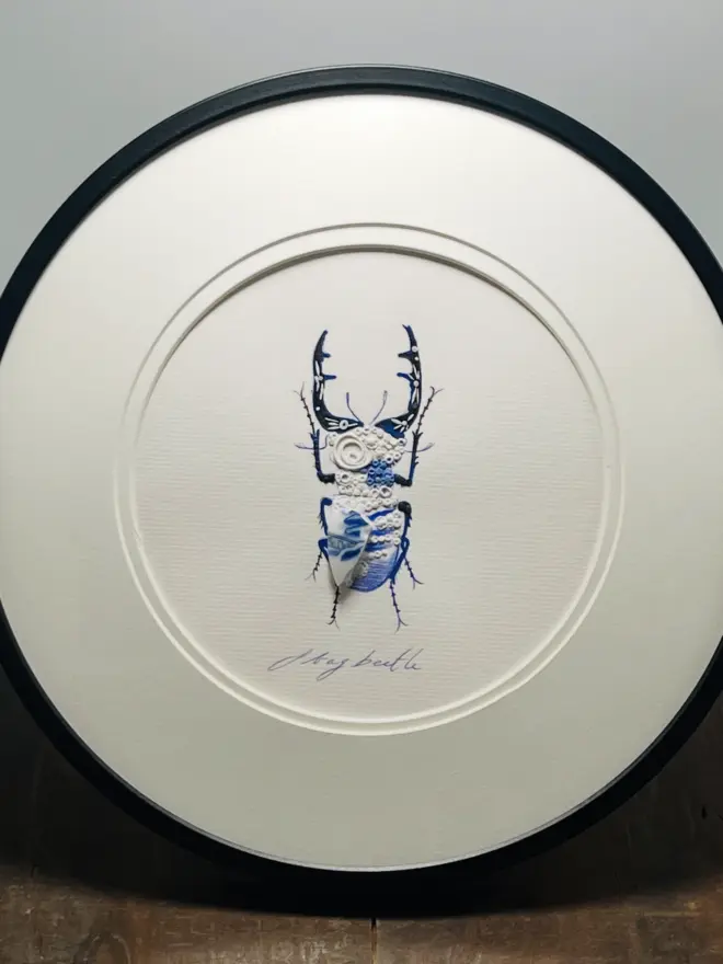 Hand-drawn, painted, mosaic clay, blue Willow Tree pottery insect on white double mounted card, in a black circular frame.