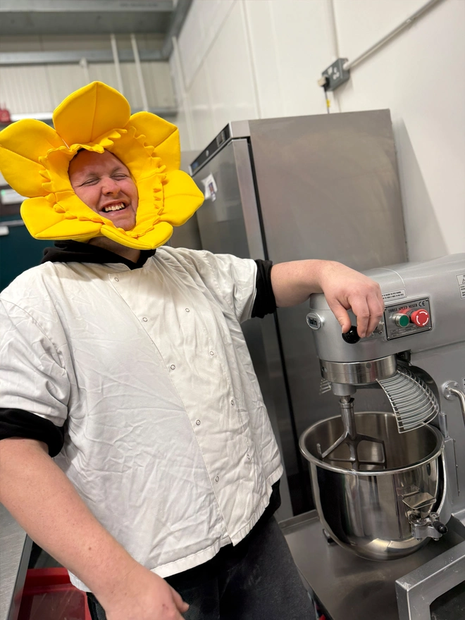 A male baker in bakers white standing next to a mixer laughing whilst wearing a daffodil hat