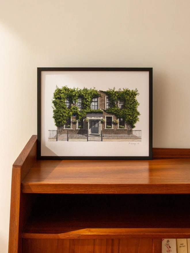 A framed watercolor print of The Albion displayed on a mid-century wooden cabinet.