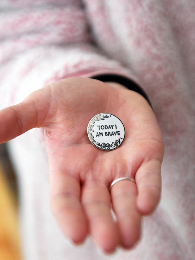 A round white enamel pin with a floral design and the words "Today I Am Brave" rests on the palm of a hand.
