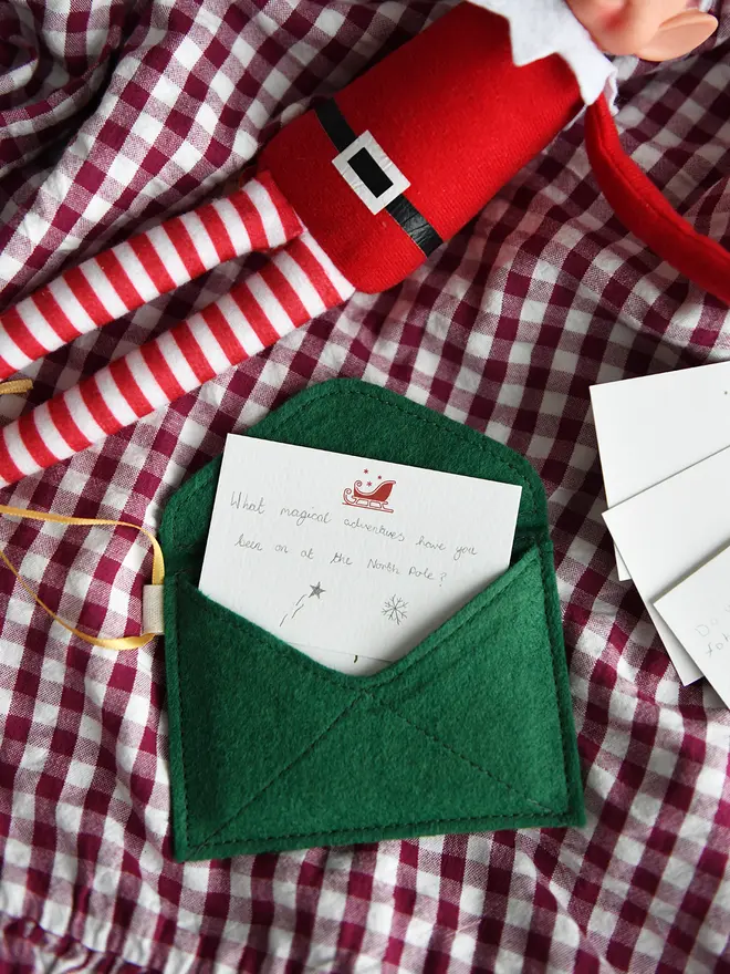 A handmade green Christmas envelope with a festive card tucked inside, written with a note to the elves, lays on a gingham cloth next to a Christmas elf toy.