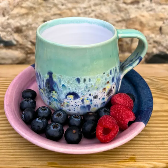Shoreline Large Round Mug, a colourful mug on a wooden surface against a brick backdrop. It is accompanied by fruits. 