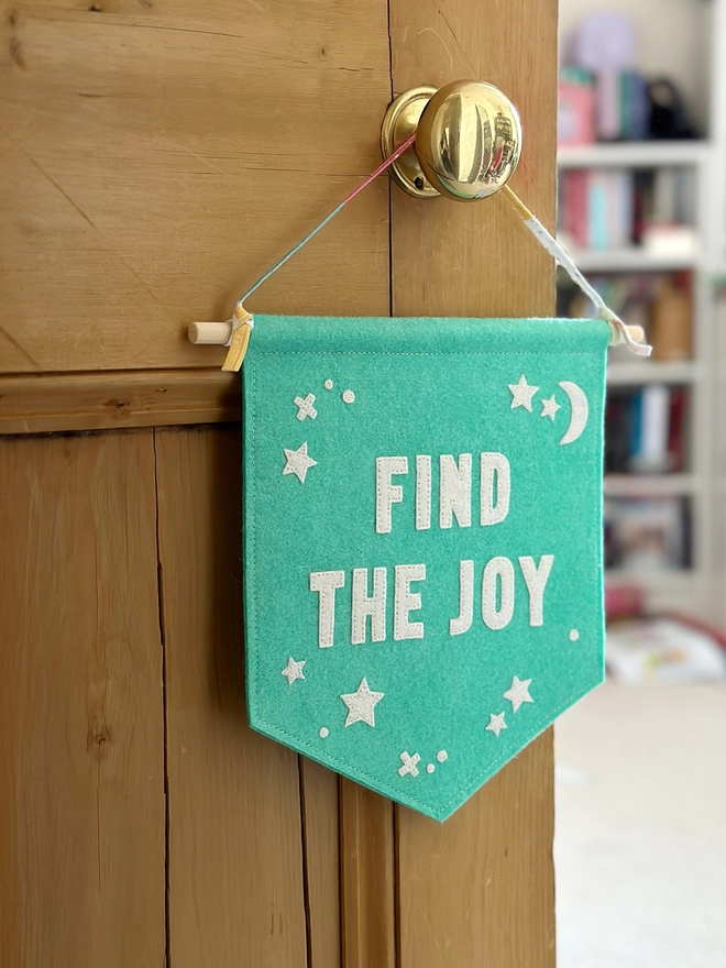 A turquoise felt banner with white words that read "Find The Joy" stitched on hangs from a ribbon hanger on a wooden door.