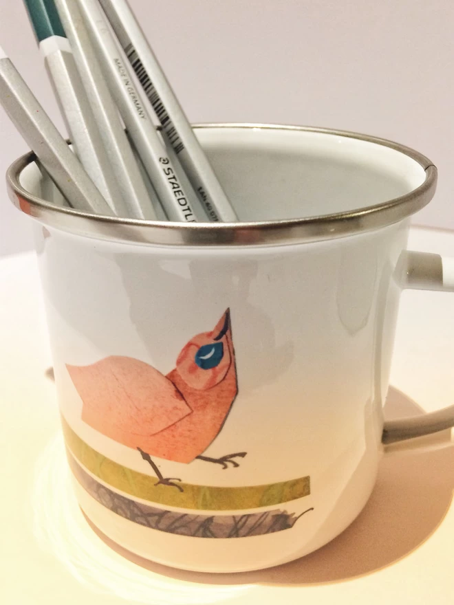 White enamel mug showing pink bird decoration. Mug holds pencils