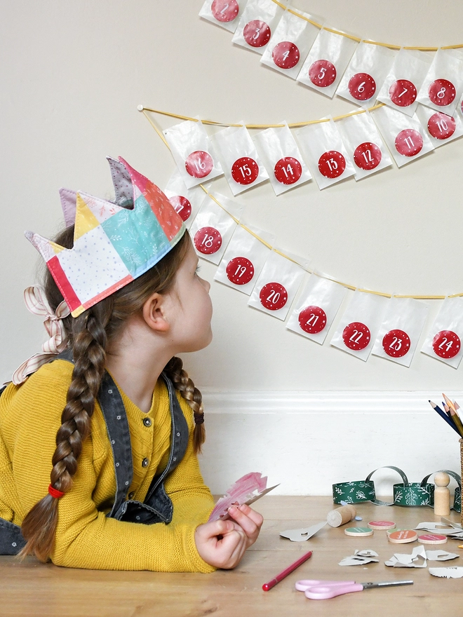 A young girl wearing a yellow top and grey dungarees, and a patchwork crown, lays on a wooden floor and looks at an advent calendar hung on the wall.