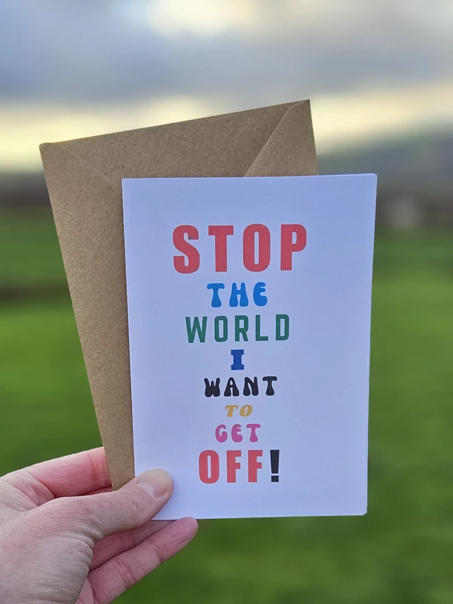 a white card with colourful writing that says stop the world I want to get off in different font styles held by a hand against a backdrop of the countryside