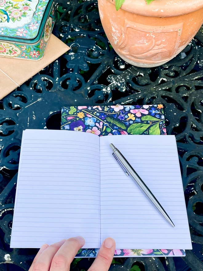 Pen on Open Lined A6 Notebook, Another Nature-Inspired Notebook with Floral Cover, on Garden Table with Plant Pot and Vintage Tin