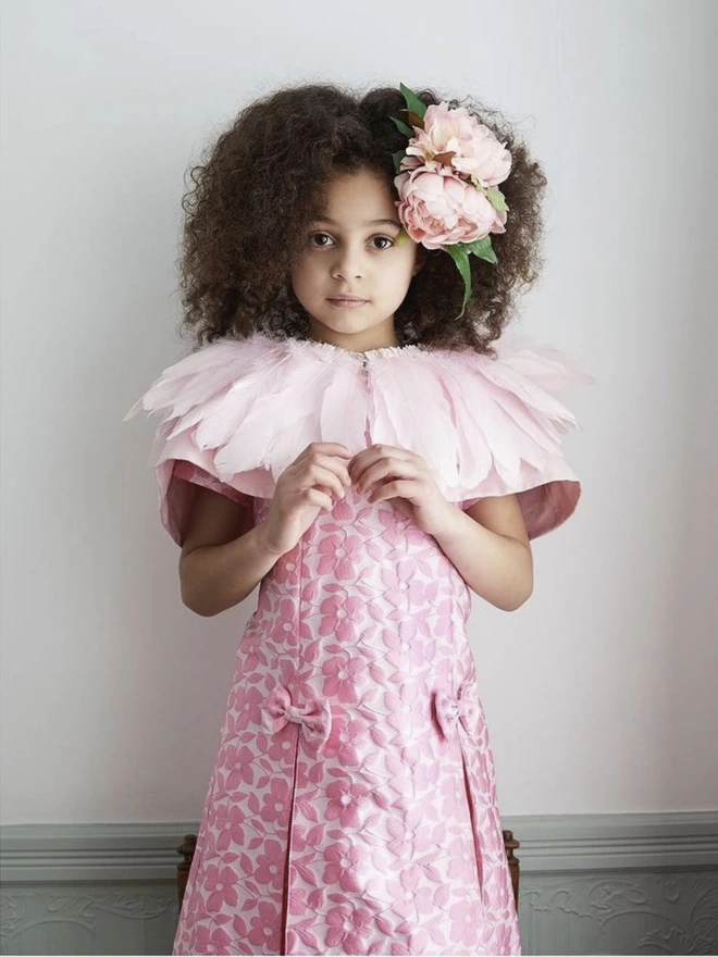 A girl standing forward with her hands in front of her chest, wearing a pink floral dress, a pastel pink feather cape and pink peonies in her curly hair