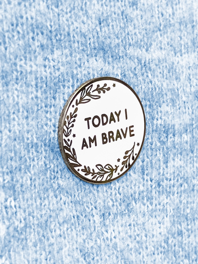 A round white enamel pin with a floral design and the words "Today I Am Brave" is pinned to blue fabric.