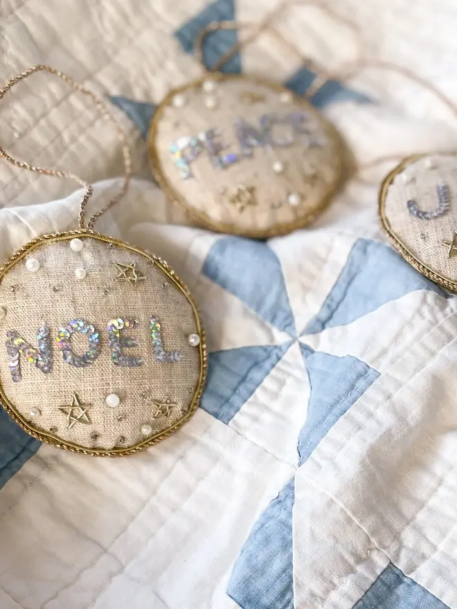embroidered linen peace joy and noel decorations. beige linen hanging decorations laid on blue and white quilt.