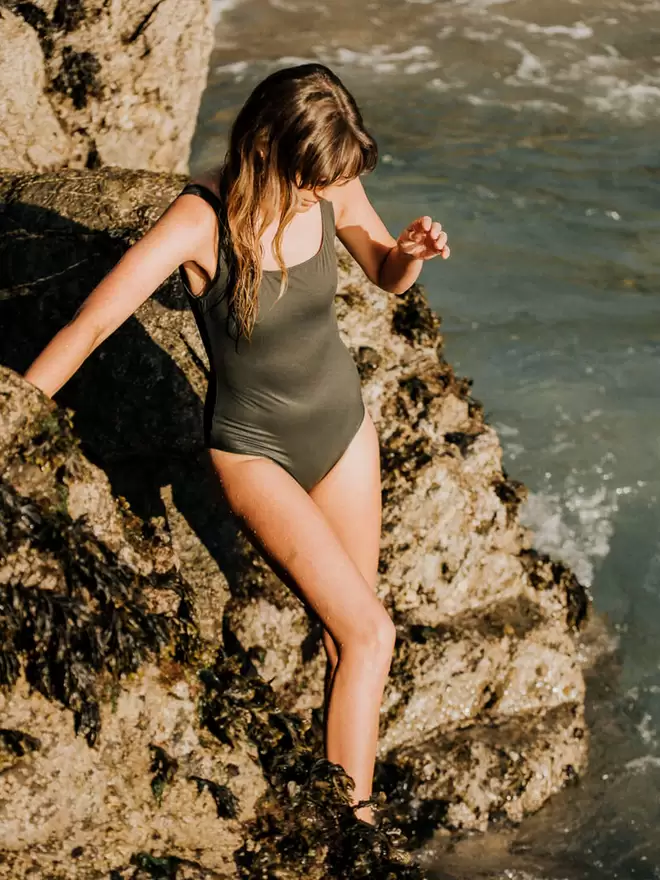 Woman with long wavy hair climbing down rocks into the sea wearing Davy J Sustainable Waterwear olive classic crossback swimsuit