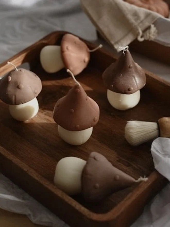 Pair Of Little Mushroom Candles (Set Of 2). A set of mushroom-shaped candles with brown dotted caps and cream stems, arranged on a wooden tray beside a linen pouch and a soft brush. The whimsical sculptural candles bring a cosy woodland feel to autumn décor.