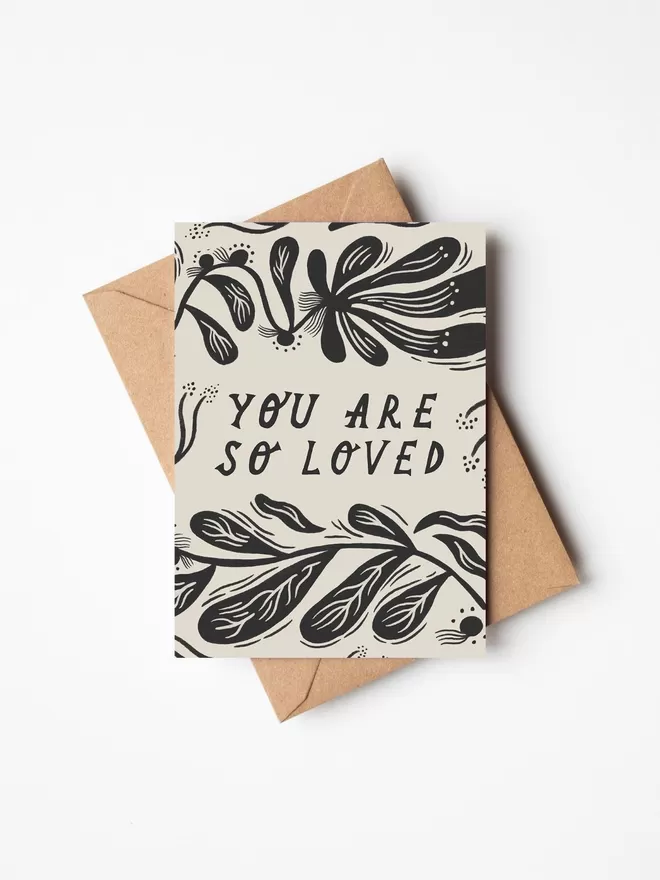 Black and white greeting card with illustration and the words you are so loved on it with a brown envelope underneath it. 