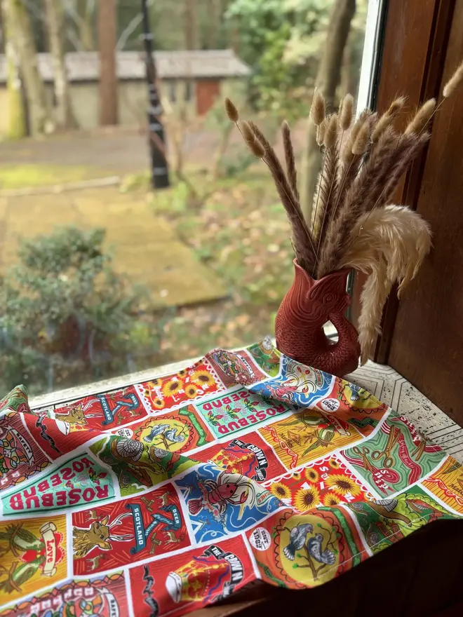 Matchbox & Tin's Print Tea Towel, a colourful decorated tea towel sitting on a window sill. 