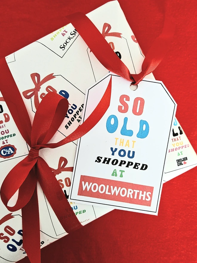 A rectangular present gift wrapped in a design that says so old that you shopped at Woolworths, C&A, Sock Shop, Tie Rack, Athena on an illustrated white tag with a red bow. The text is written in bright  multi colours .