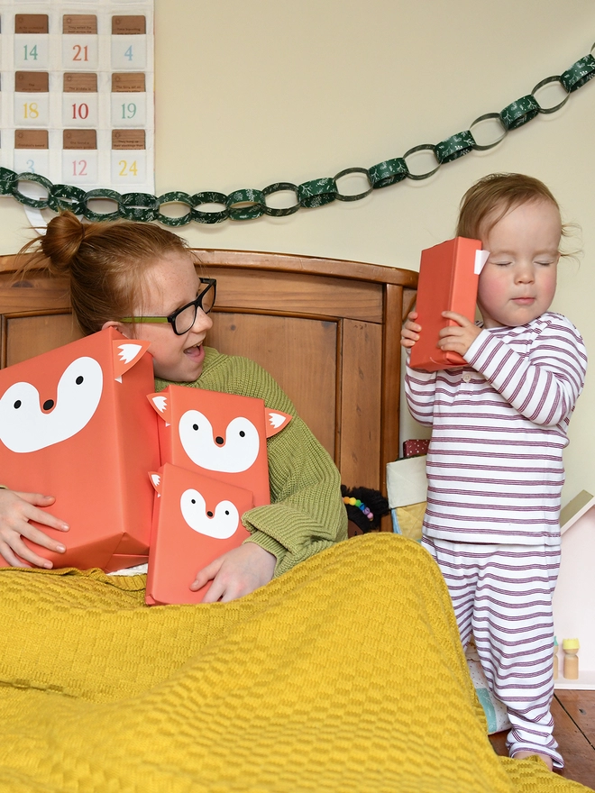 Two children sat in a wooden bed hold gifts wrapped in orange fox wrapping paper.