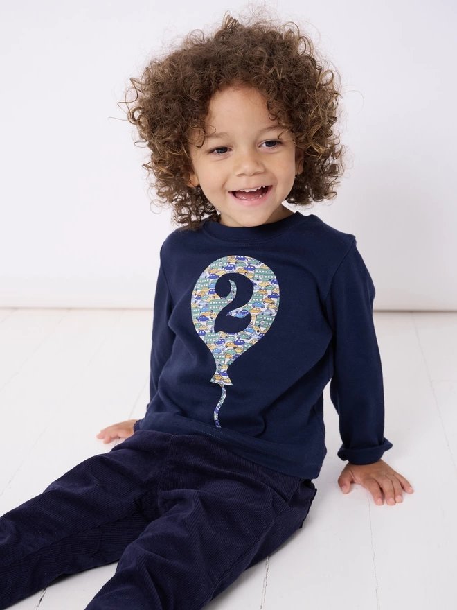 A smiling 2 year old boy wearing a navy cotton long sleeve t-shirt. The t-shirt features a balloon with the number 2 cut out from it, appliquéd in a cars Liberty print.