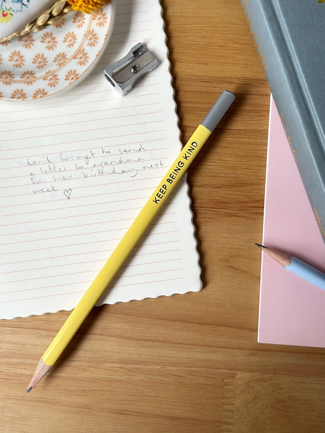 A yellow pencil with the words Keep Being Kind along the side in silver lettering lays on an open lined notebook on a wooden desk. Various stationery items are beside it.