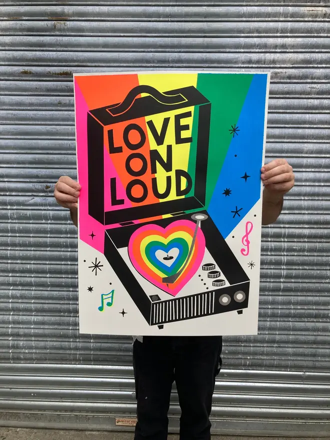 'Love On Loud' Screenprint, a person holding a colourful screenprint in front of a metal shutter. The screenprint says 'Love on loud'. 