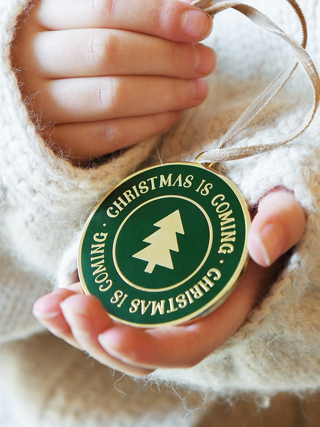 A deep green and gold enamel Christmas decoration, with the words Christmas Is Coming surrounding a gold Christmas tree, is being held by two hands.