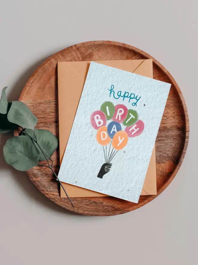 Plantable Card with an an illustrated hand holding balloons with a letter in each balloon spelling out Happy birthday on a wooden tray next to a Eucalyptus branch