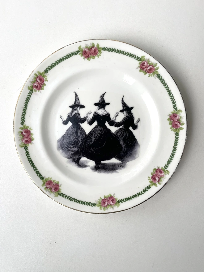 a coven of 3 witches dance hand in hand, the image is victorian in black and white and is printed directly onto an ornate vintage plate