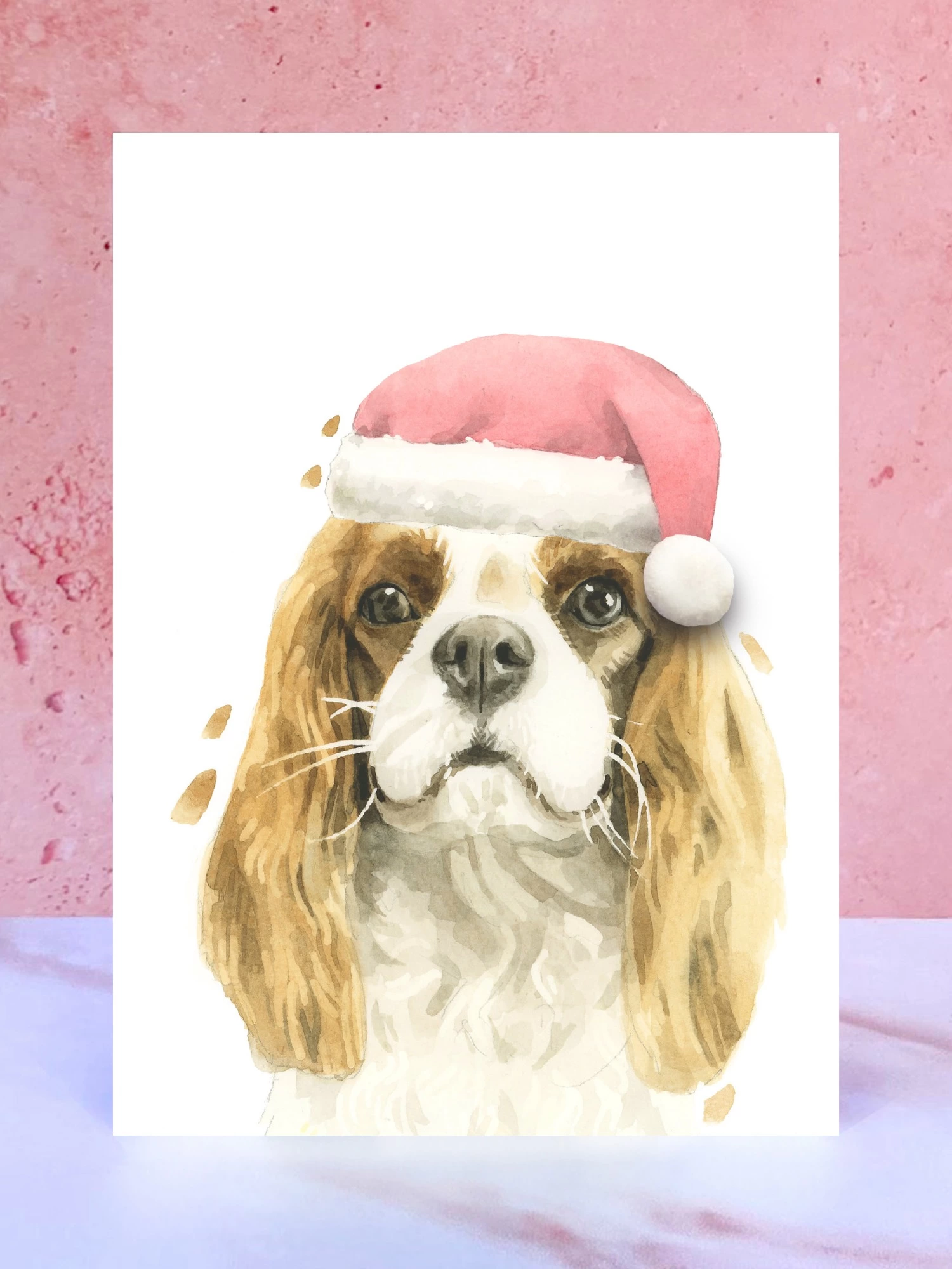 A Christmas card featuring a hand painted design of a cavalier king charles spaniel, stood upright on a marble surface surrounded by pompoms. 