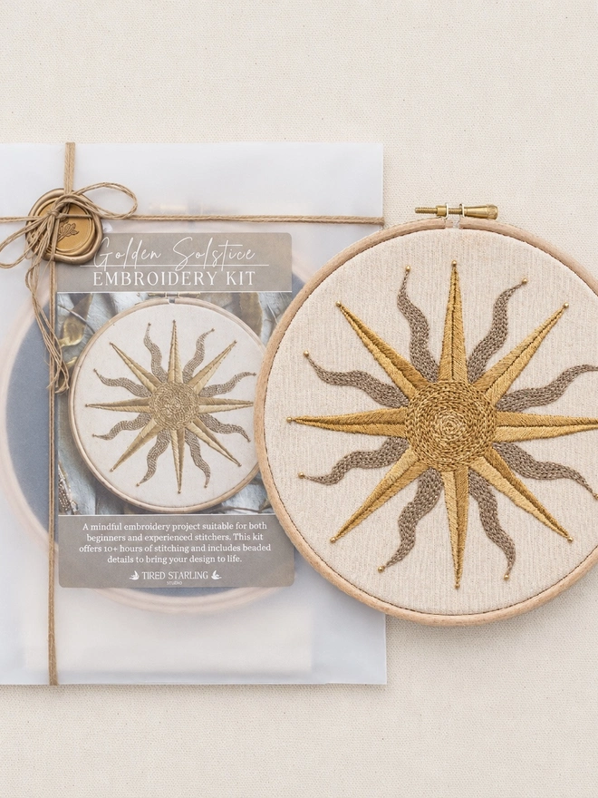 Golden Solstice embroidery kit packaging displayed beside the finished embroidered sun hoop. The kit is wrapped in translucent paper and tied with twine, featuring a celestial sun design in warm neutral tones.