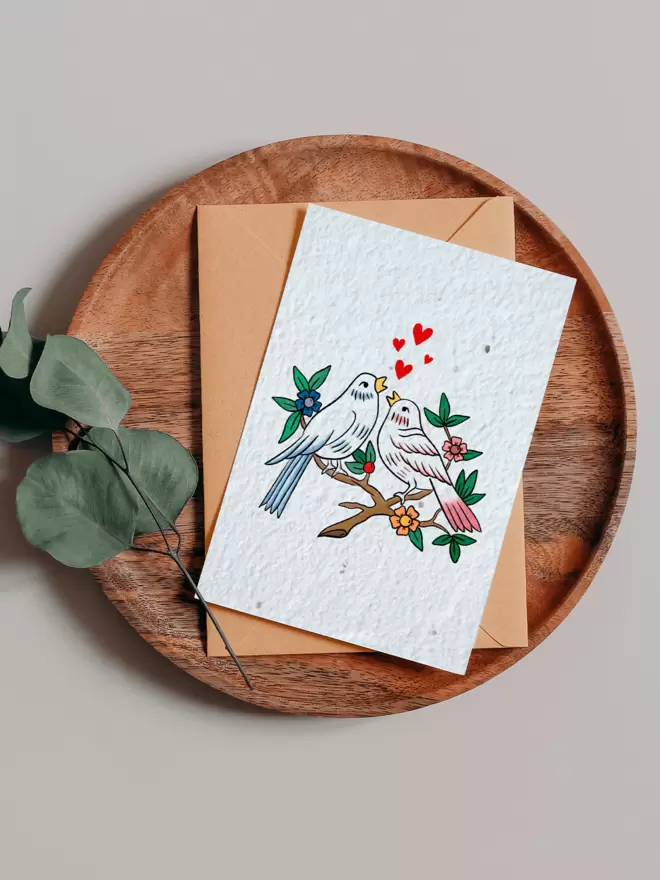 Seeded Paper Greeting Card featuring an illustration of two birds sitting on a branch in the centre on a wooden tray next to a Eucalyptus branch