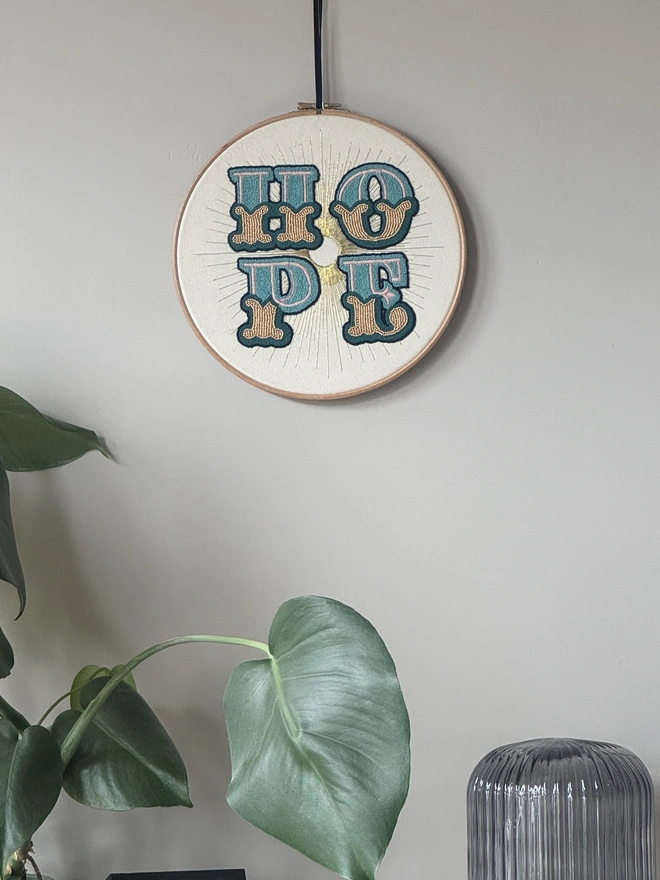 An embroidery hoop hanging on a wall containing the stitched word Hope in green and gold 