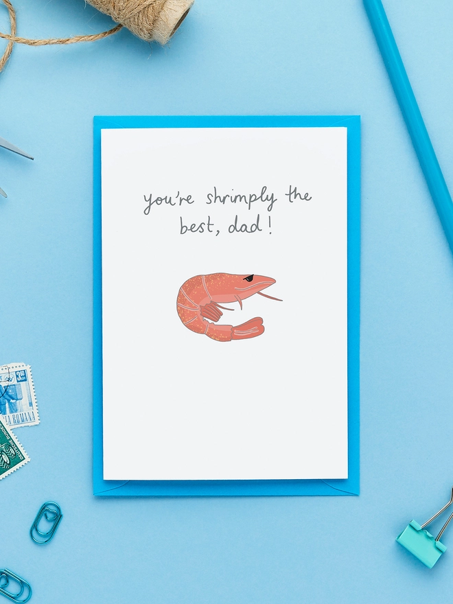 Greeting card with a hand-drawn shrimp and the caption “You’re shrimply the best, dad!”, styled on a blue background with twine, pencil, stamps and envelope.