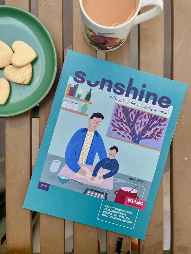 Sonshine magazine issue 21 a green mag with an illustration of father and son baking on the cover on a garden table with a cup of tea and some heart shaped biscuits 