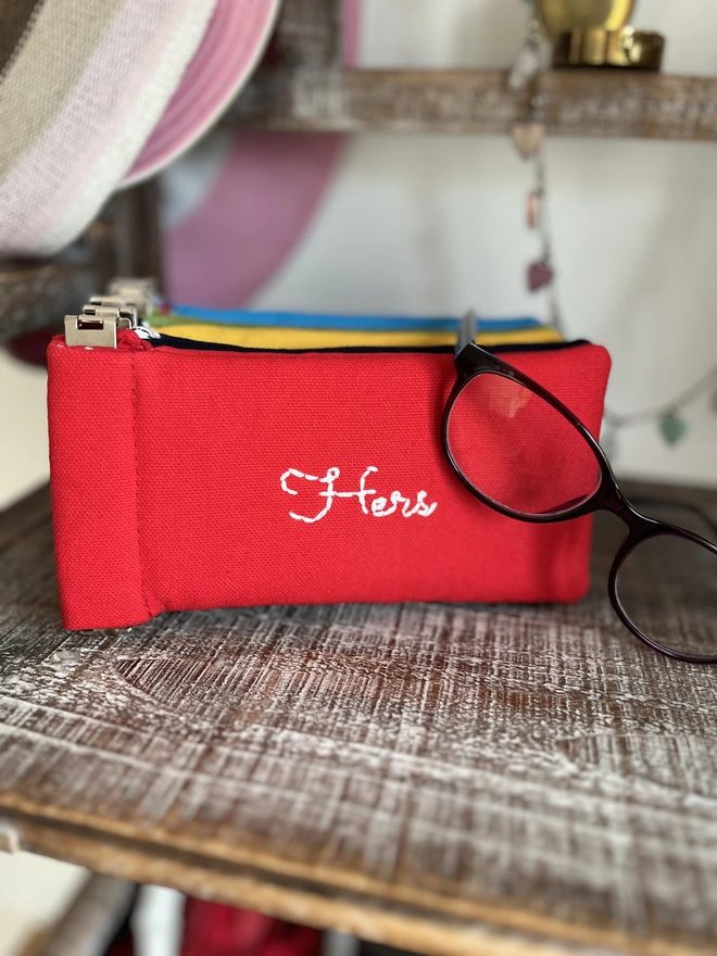 Red cotton glasses pouch embroidered with 'Hers'. Pair of glasses draped of it. Alternative bright colours in the soft case in background.