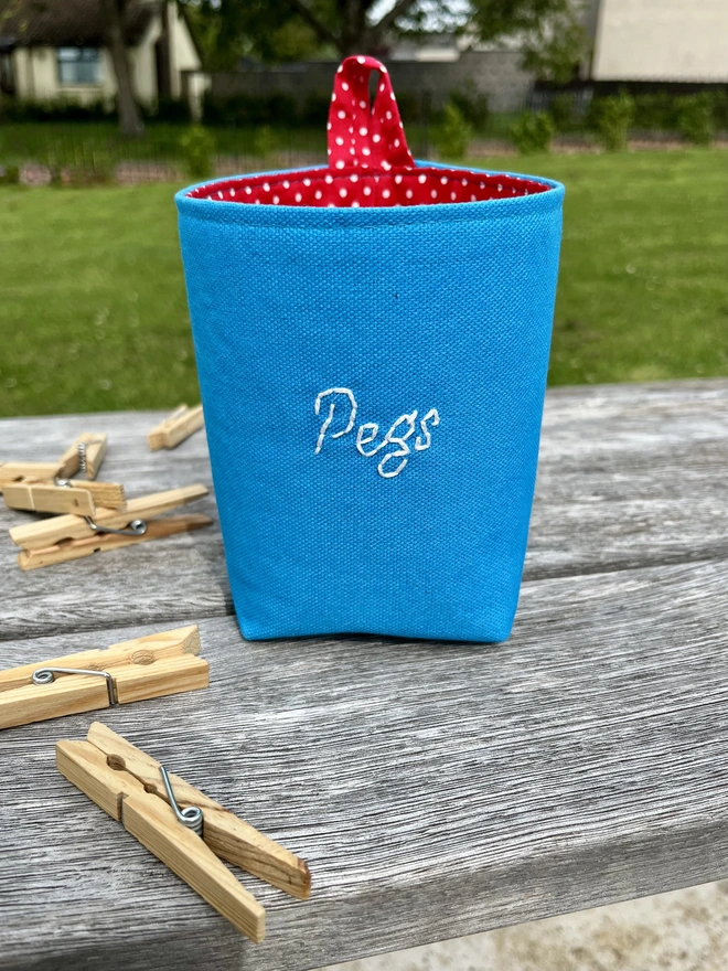 Blue fabric bag outdoors on a bench with clothes pegs around it, embroidered with ‘Pegs’ on front