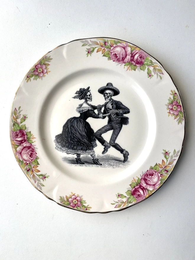 vintage plate with an ornate border, with a printed vintage illustration of two dancing day of the dead skeletons wearing a dress and a suit in the middle