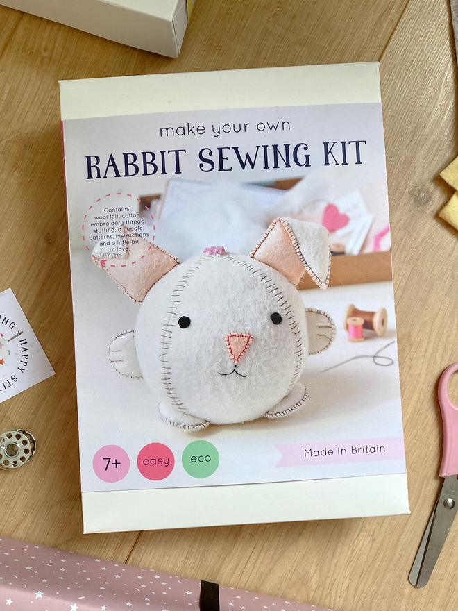 A craft kit box with an image of a white felt rabbit toy and the words "Make your own rabbit sewing kit" lays on a wooden desk. Various craft kit materials lay beside the box.