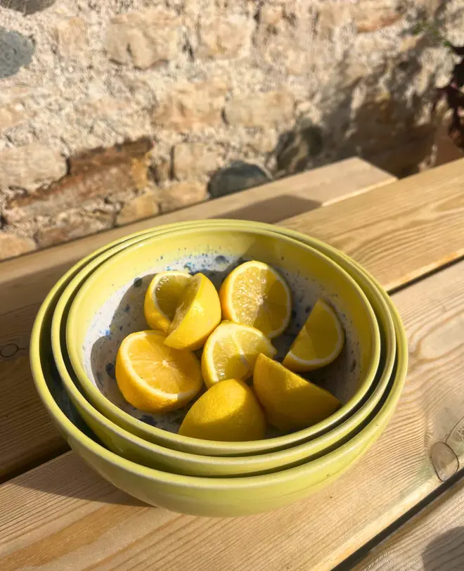 Shoreline Serving Bowls (Set Of 3), a collection of colourful serving bowls on a wooden surface against a brick backdrop. They are accompanied by fruits. 