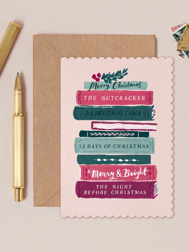 Pale pink Bookish Christmas card with a scalloped edge and a tall stack of Christmas story books, on a kraft envelope beside a gold pen