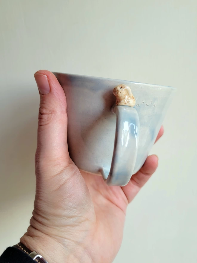 A shiney grey glazed handmade pottery  coffee cup with a little beige bunny rabbit and footprints held in a hand