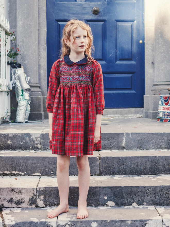 A girl stands on stone steps wearing a red tartan dress with smocking and a navy collar
