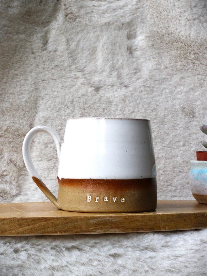 Rustic ceramic mug with a white speckled upper glaze and brown lower half, featuring the word “Brave” on the front, sitting on a wooden table against a neutral background.