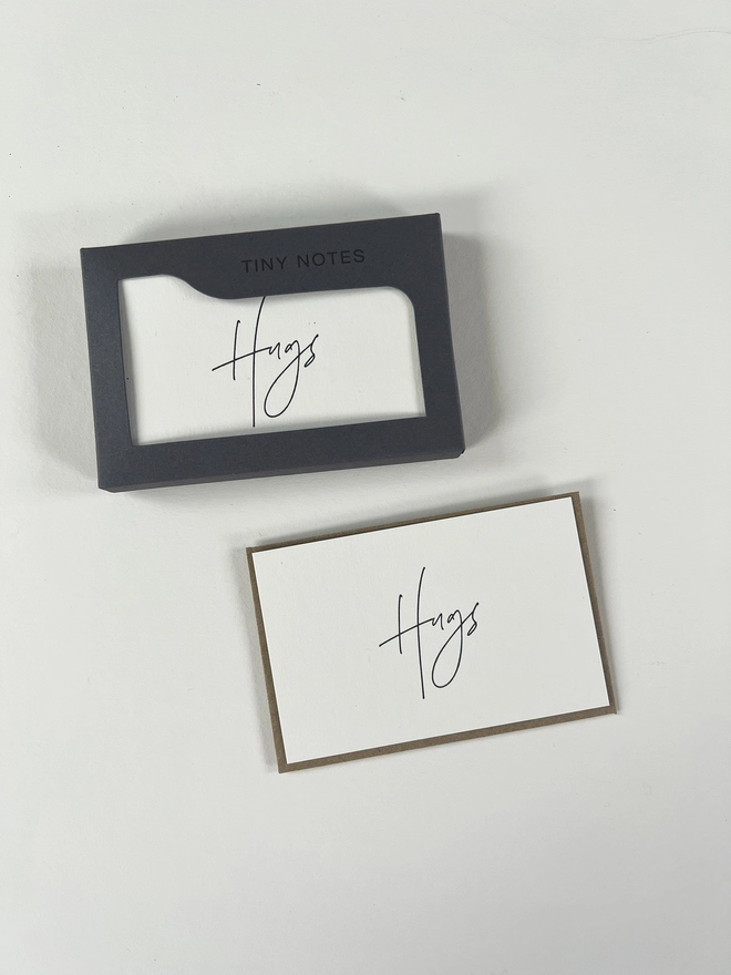 Letterpress hugs notes in their letterpress box, image shows one card out of the box with it's Kraft brown recycled envelope. Hugs is written in a modern calligraphy style.