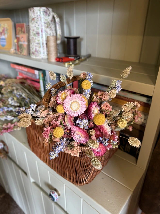 a bouquet of dried flowers in bright colours displayed in a wicker basket against a green painted dresser
