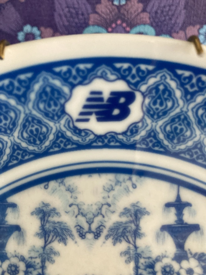 detail of plate showing the New Balance logo. The plate is blue and white in a Delft style featuring an illustration of a New Balance trainer in the centre