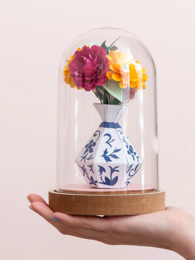 A hand holding a paper sculpture that features a bunch of paper flowers in a white and blue vase on a plastic dome . This is a craft kit by My papercut Forest