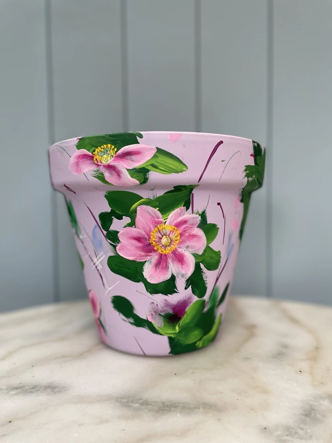 A beautifully hand painted interior terracotta plant pot. Background painted in a pale dusky pink adorned in pink Japanese anemone. 